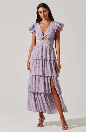 A woman is wearing a light purple, floral-patterned dress with multiple tiered ruffles. The dress features short, puffed sleeves with ruffled details, a deep V-neckline, and a cut-out detail at the waist. The skirt has a high slit on the left side, revealing her leg. She is also wearing strappy high-heeled sandals.