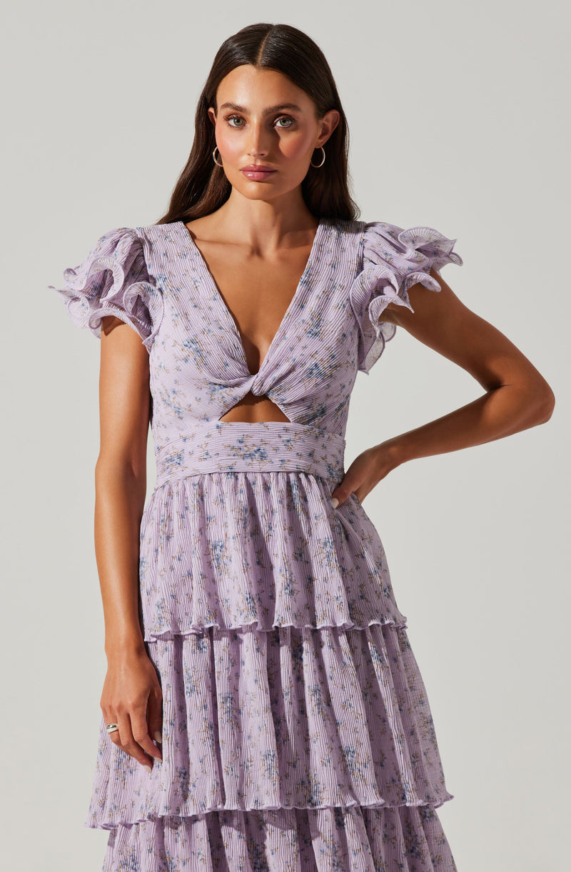 A woman is wearing a lavender-colored dress with a floral pattern. The dress features a deep V neckline with a twist knot detail at the center, creating a small cutout below the bust. It has short, ruffled sleeves, a fitted waist, and a tiered, ruffled skirt.