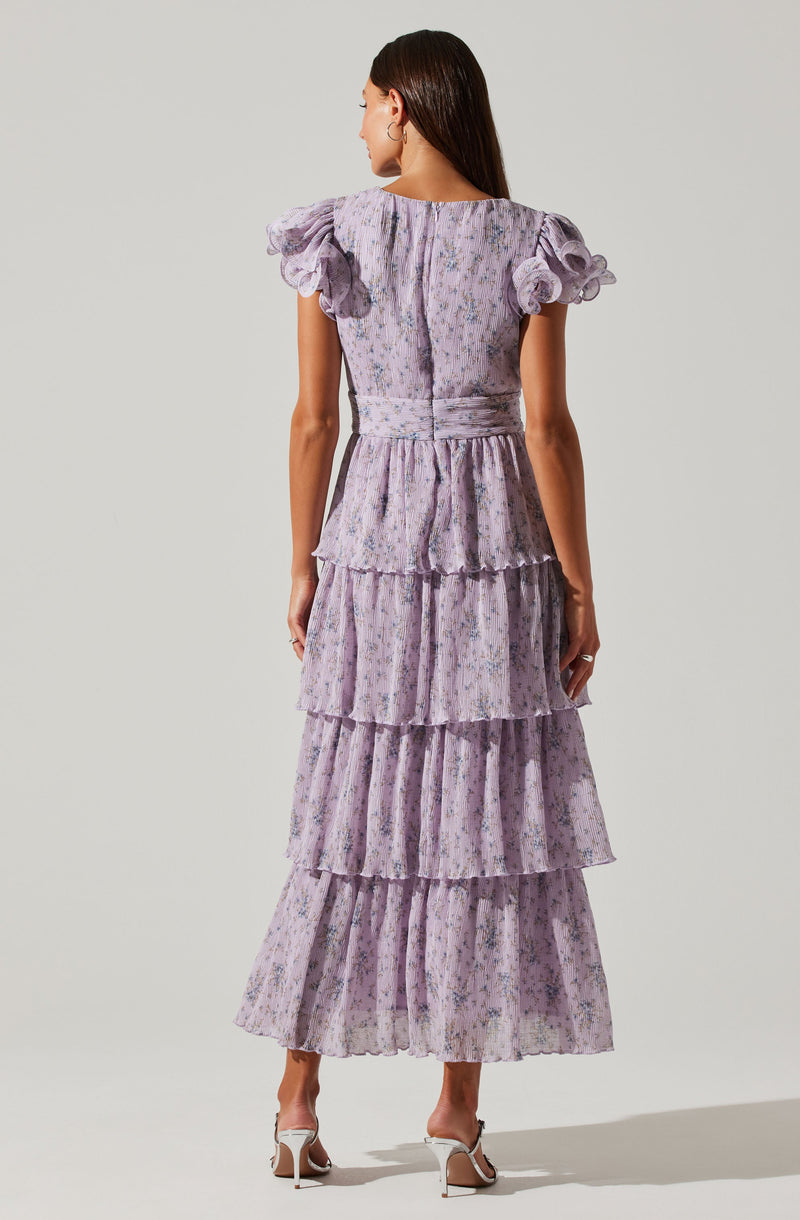 A woman is wearing a long, lavender-colored dress with a floral pattern. The dress features ruffled, puffed cap sleeves and a fitted waist. It has multiple tiered ruffles that extend from the waist to the hem, creating a layered, flowing look. The fabric appears lightweight and textured. She is also wearing white high-heeled sandals.