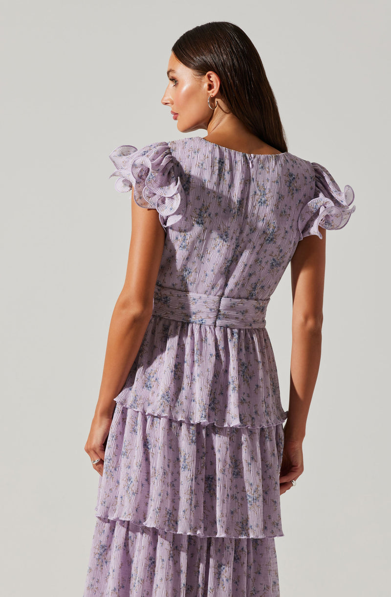 A woman is wearing a light purple dress with a floral pattern. The dress features ruffled, gathered cap sleeves with multiple layers. The fabric is textured with a subtle vertical stripe pattern, and the waist is cinched, creating a fitted silhouette. The skirt has tiered ruffles that flow down to the hem.