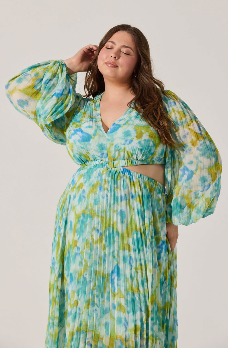 A woman is wearing a matching two-piece outfit with a colorful, abstract pattern in shades of blue, green, and yellow. The top has a V-neckline, long puffed sleeves, and a cut-out at the midriff. The skirt is high-waisted with a pleated design.