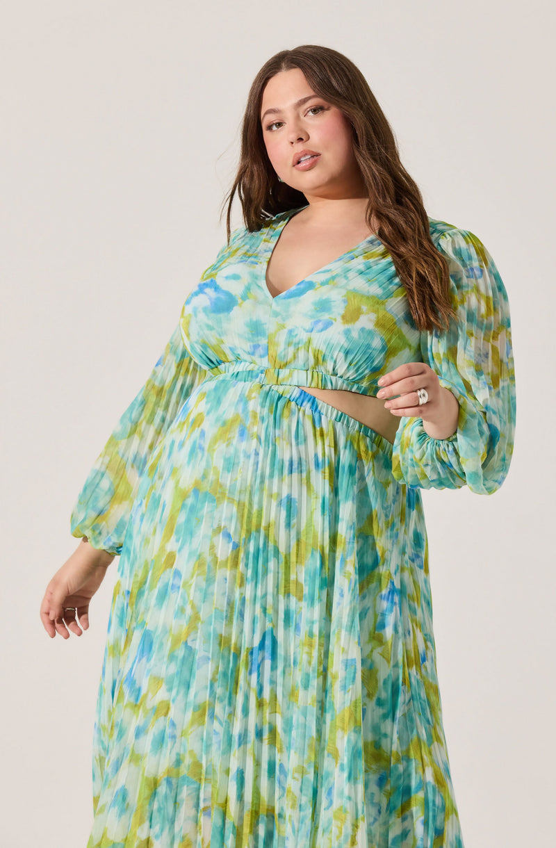 A woman is wearing a long, flowing dress with a multicolored, watercolor-style pattern in shades of blue, green, and yellow. The dress has long, puffed sleeves with elastic cuffs, a V-neckline, and a cutout at the midriff.