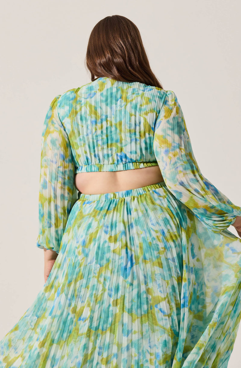 A woman is wearing a matching set of a light, airy, tie-dye print blouse and skirt in shades of blue, green, and yellow. The blouse features long, slightly puffed sleeves and a cropped style with an elasticized hem. The skirt is high-waisted with pleats, creating a flowing silhouette. The fabric appears lightweight and semi-transparent.