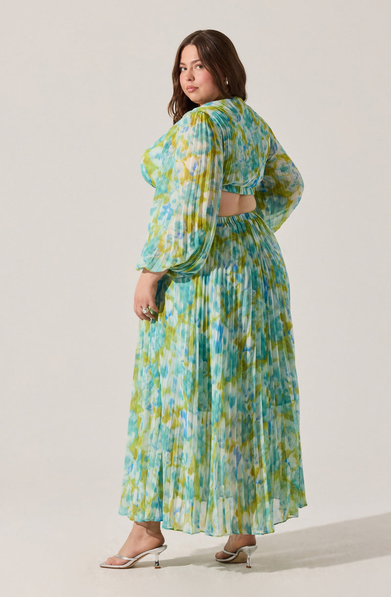 A woman is wearing a flowy, two-piece outfit with a multicolored abstract pattern in shades of blue, green, and yellow. The top has long, puffed sleeves and a cut-out detail at the back, revealing her midriff. The skirt is high-waisted, pleated, and reaches down to her ankles. She is also wearing light-colored, open-toe heels.