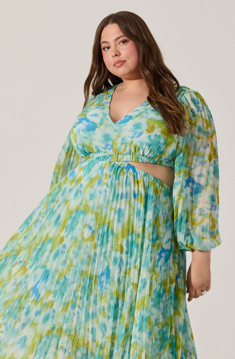 A woman is wearing a long, flowing dress with a multicolored abstract pattern in shades of blue, green, and white. The dress features a v-neckline, puffed three-quarter sleeves, and a cut-out detail at the waist.