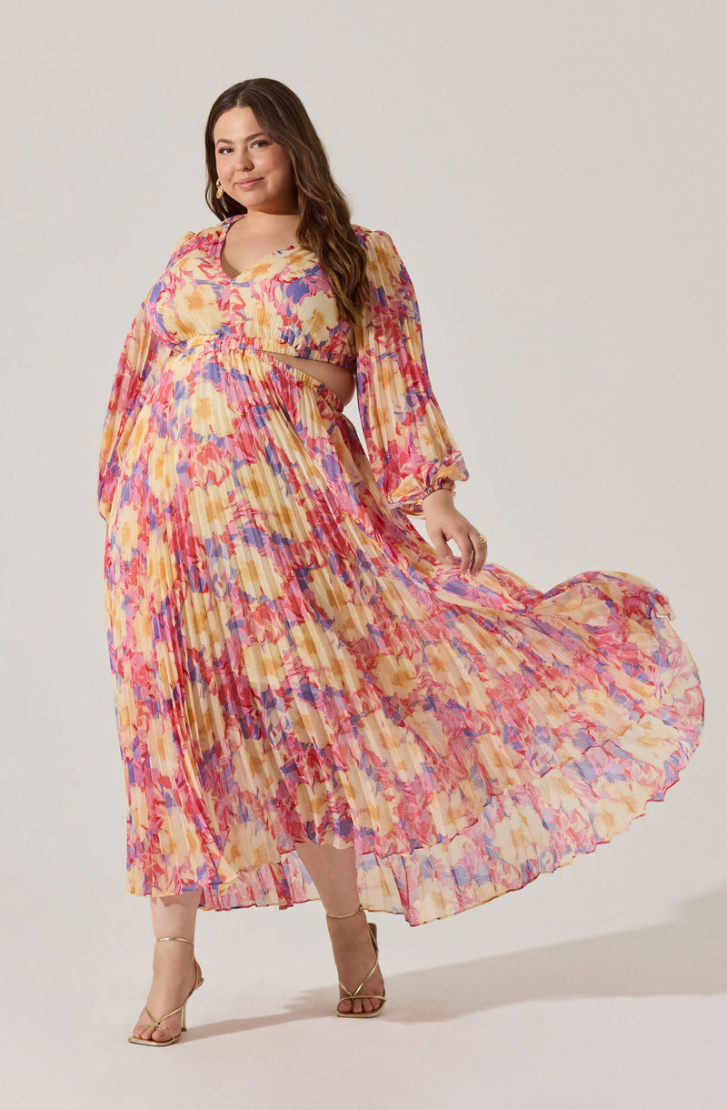 A woman is wearing a colorful, flowing maxi dress with a vibrant floral pattern in shades of pink, yellow, purple, and orange. The dress features long, slightly puffed sleeves with elastic cuffs and a gathered bodice with a cutout in the midriff area. She pairs it with beige high-heeled sandals. The overall look is bright and summery.