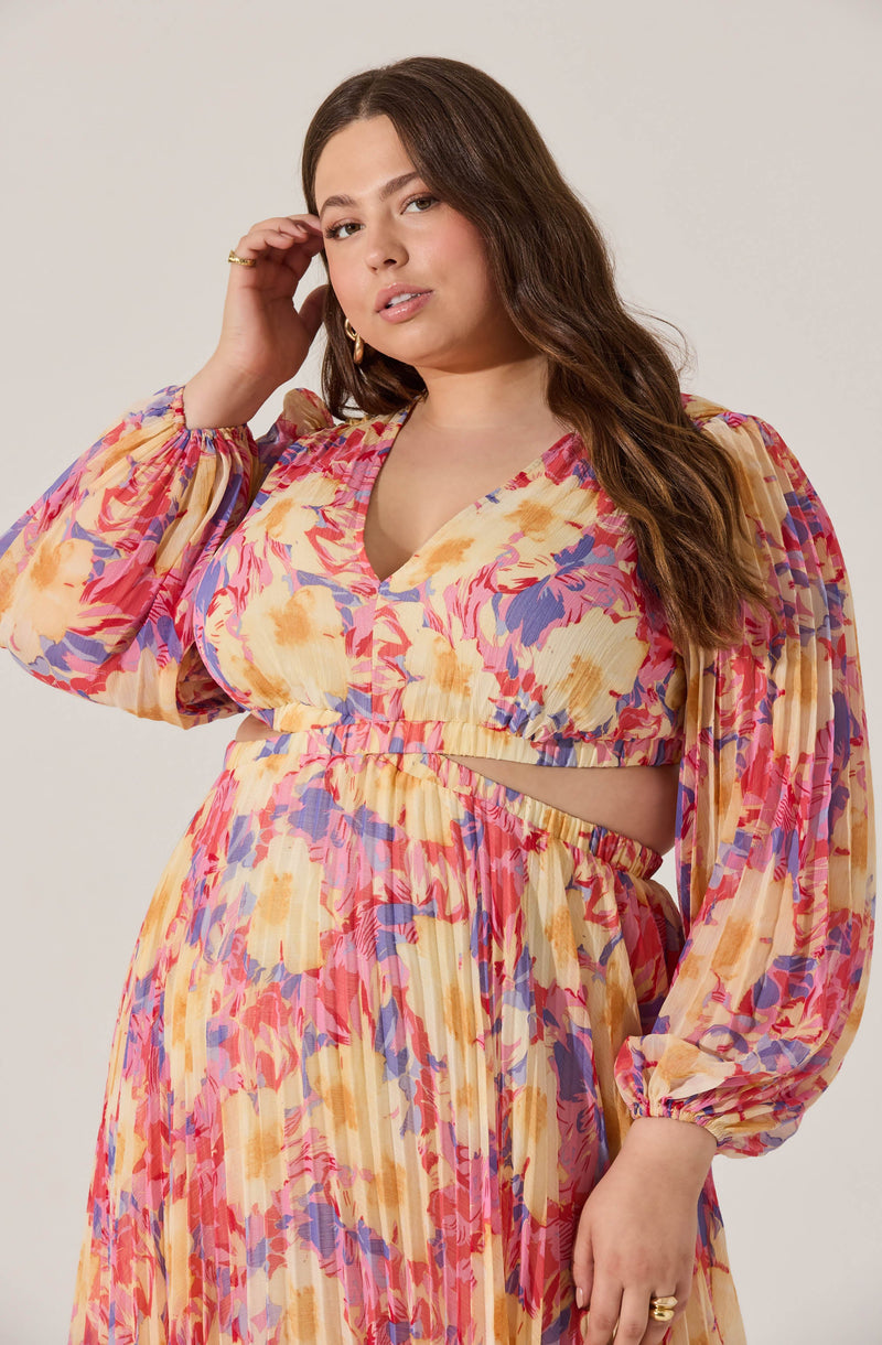 A woman is wearing a colorful, floral-patterned outfit that includes a long-sleeved, cropped top with puffed sleeves and a V-neckline, paired with a matching high-waisted skirt. The top has elastic at the waist creating a gathered look, and the skirt features an elastic waistband. The fabric of both garments features a vibrant, abstract floral print in shades of pink, yellow, purple, and peach.