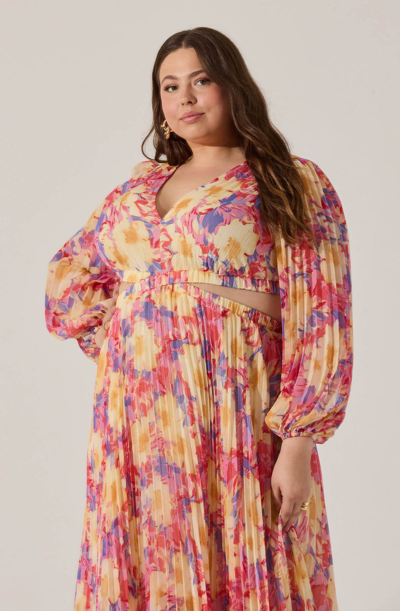 A woman wearing a colorful, floral-patterned outfit consisting of a long flowing skirt and a matching blouse. The blouse features long, puffed sleeves and a deep neckline, with a cut-out detail above the waist. The fabric has a vibrant mix of yellow, pink, purple, and beige hues arranged in an abstract floral design.