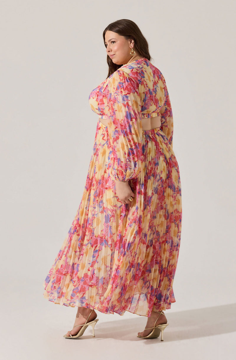 A woman is wearing a long, flowy dress with a colorful, abstract pattern in shades of pink, purple, orange, and yellow. The dress has long, puffed sleeves, a high neckline, and a cutout at the back with a beige band detail. She is also wearing beige high-heeled sandals and gold hoop earrings.