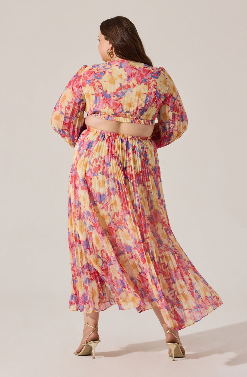 A woman is wearing a two-piece outfit with a colorful, floral pattern. The top features long, puffed sleeves and a cropped length, exposing the midriff. The skirt is a high-waisted, ankle-length, flowy maxi with a matching vibrant, abstract floral print. The fabric appears lightweight and semi-sheer with pleats. She pairs the outfit with nude high-heeled sandals.