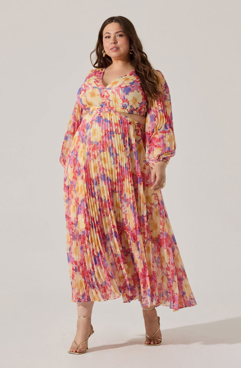 A woman is wearing a long, flowy, pleated dress with a vibrant, abstract floral pattern in shades of pink, purple, yellow, and beige. The dress has long, billowy sleeves with elastic cuffs and a matching fabric belt at the waist. The top part features a V-neckline. She is also wearing open-toe, ankle-strap heels in a neutral tone.