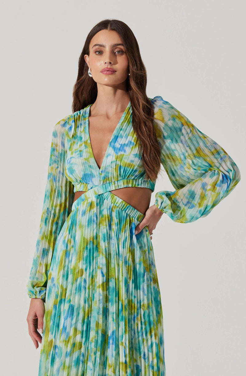 A woman is wearing a two-piece outfit with a vibrant, multicolored, watercolor-like pattern in shades of blue, green, and yellow. The top has long, semi-sheer, puffed sleeves with elastic cuffs, a V-neckline, and a cut-out detail at the midriff. The skirt is high-waisted, flowing, and pleated, matching the pattern of the top. The overall look is stylish and summery.