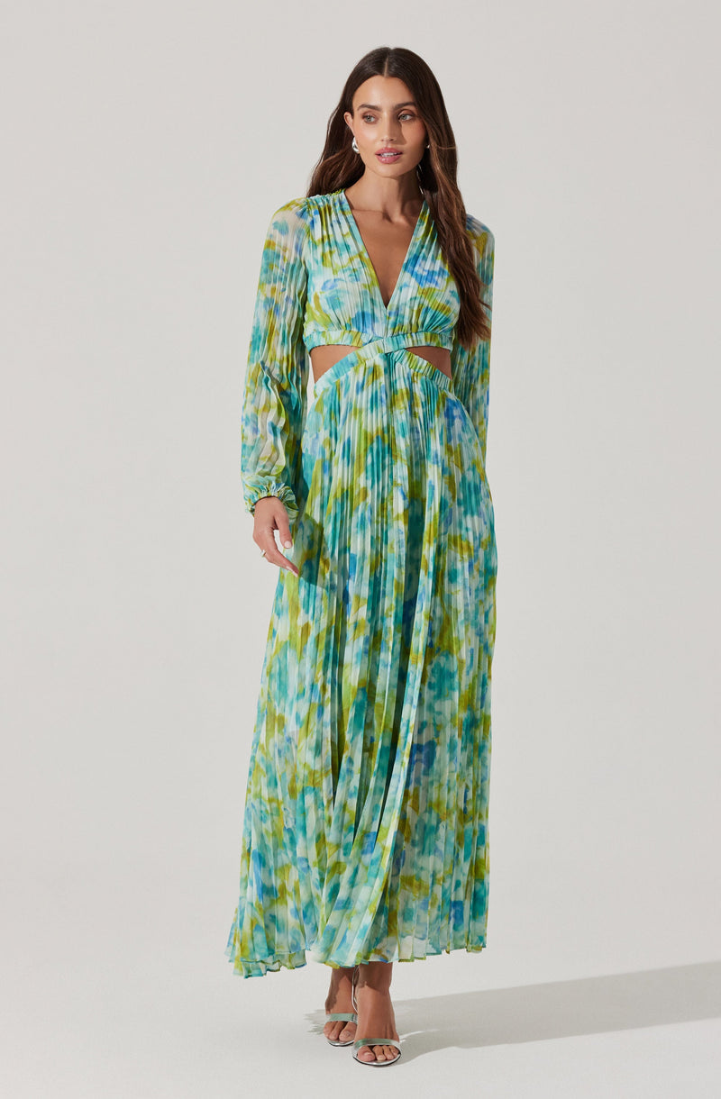 A woman is wearing a long, flowing dress with a vibrant, multicolored tie-dye pattern in shades of blue, green, and yellow. The dress features a deep V-neckline, long sheer sleeves with gathered cuffs, and cut-out details at the waist. It has pleats running vertically down the skirt, creating a lightweight, airy appearance.