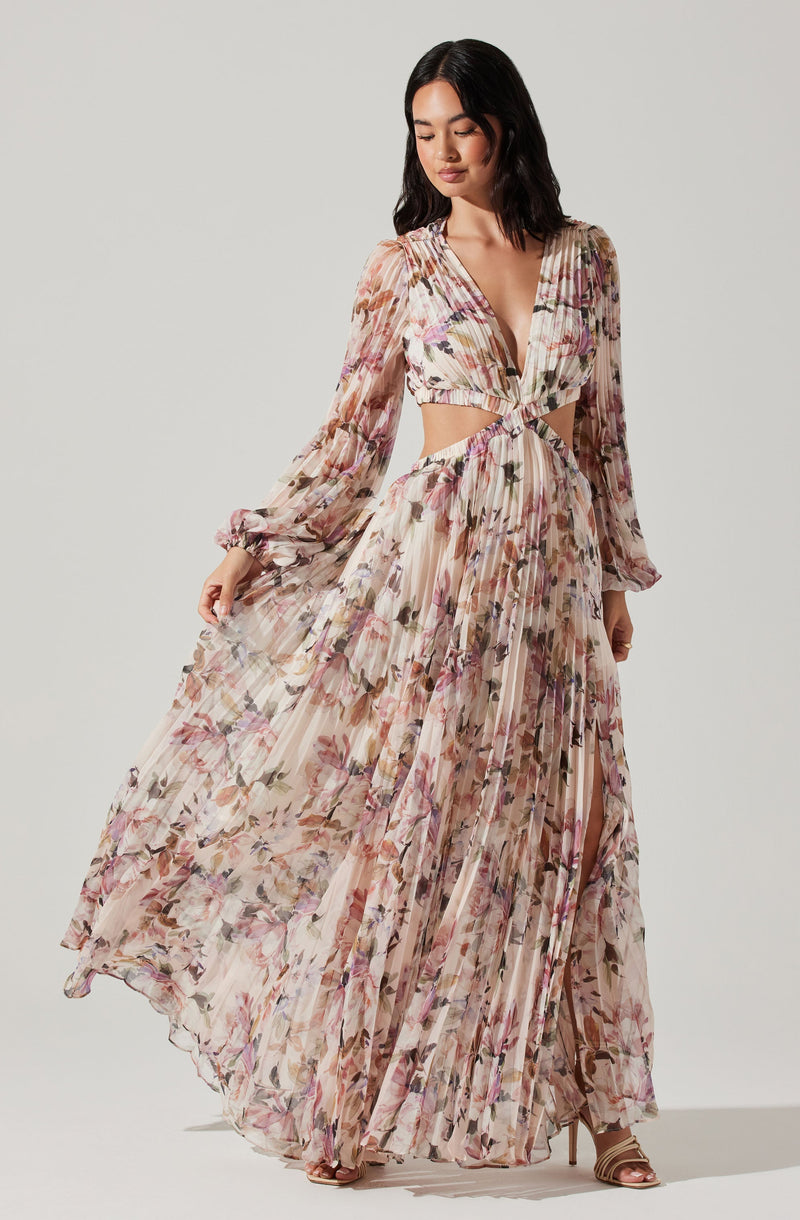 A woman is wearing a flowing, floor-length dress with a floral print in soft, pastel shades. The dress has long, semi-sheer puffed sleeves with elastic cuffs, a deep V-neckline, and cut-out sections at the waist, creating a midriff-baring effect. The skirt is pleated and semi-transparent, giving an airy appearance. She is also wearing open-toed high heels.