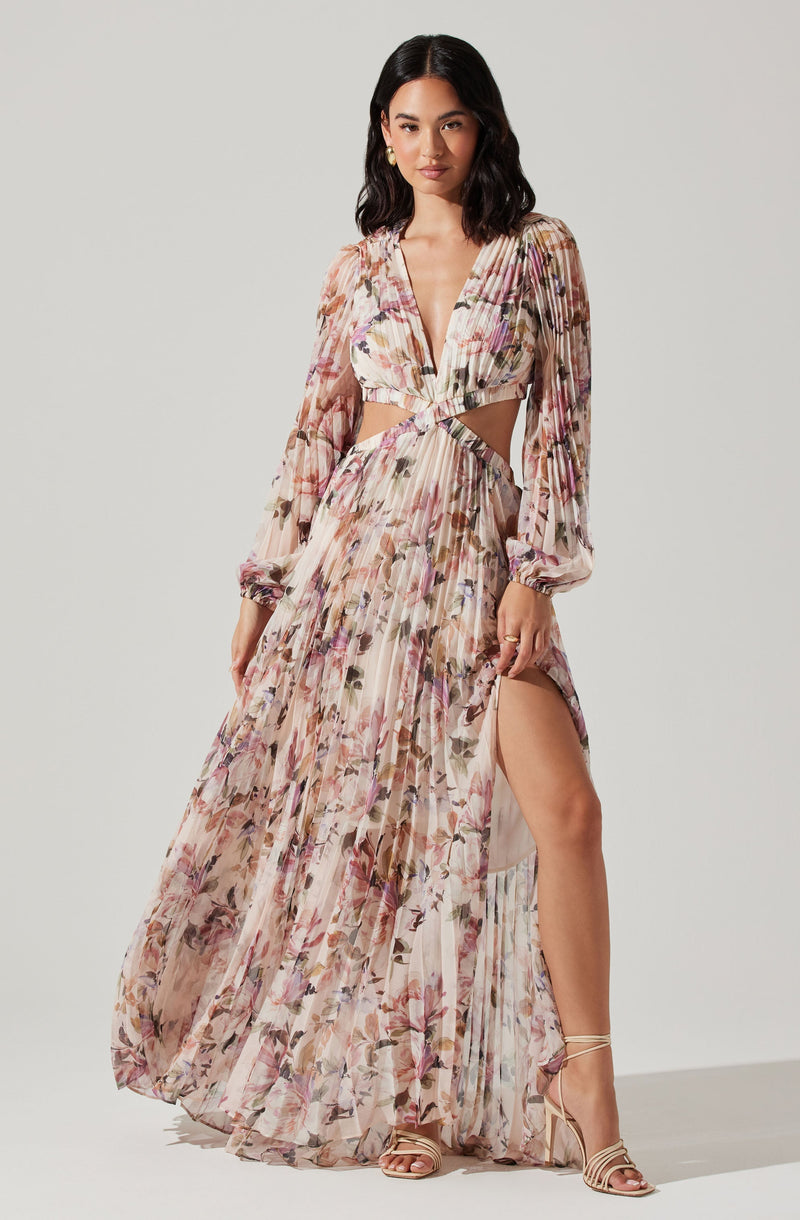 A woman is wearing a long, flowing dress made of sheer fabric with a floral pattern in shades of pink, beige, and purple. The dress features long, slightly puffed sleeves, a deep V-neck, and cut-out sections at the waist revealing skin. The skirt has a high slit on one side, showing her leg and sandals.