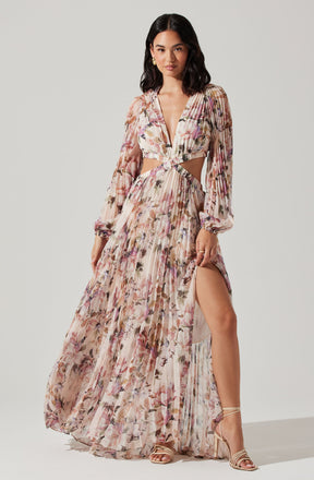 A woman is wearing a long, flowing dress made of sheer fabric with a floral pattern in shades of pink, beige, and purple. The dress features long, slightly puffed sleeves, a deep V-neck, and cut-out sections at the waist revealing skin. The skirt has a high slit on one side, showing her leg and sandals.