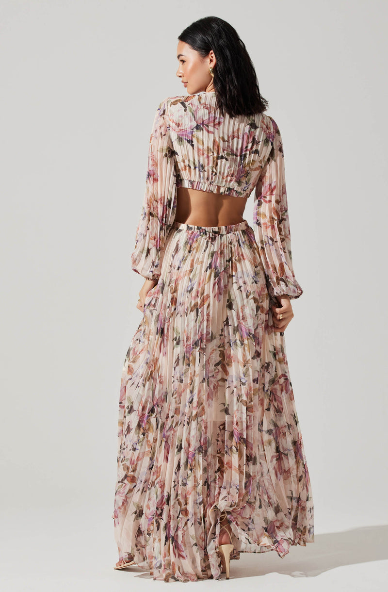 A woman is wearing a matching set composed of a long-sleeved, cropped blouse and a high-waisted, floor-length skirt. The fabric features a light, sheer material with a floral pattern in shades of pink, purple, brown, and beige, with pleats throughout. The blouse has a gathered elastic hem at the waist and puffed sleeves. The skirt is flowing and slightly transparent, with a ruffled hem at the bottom. She is standing with her back slightly turned to the camera, showcasing the outfit's details.