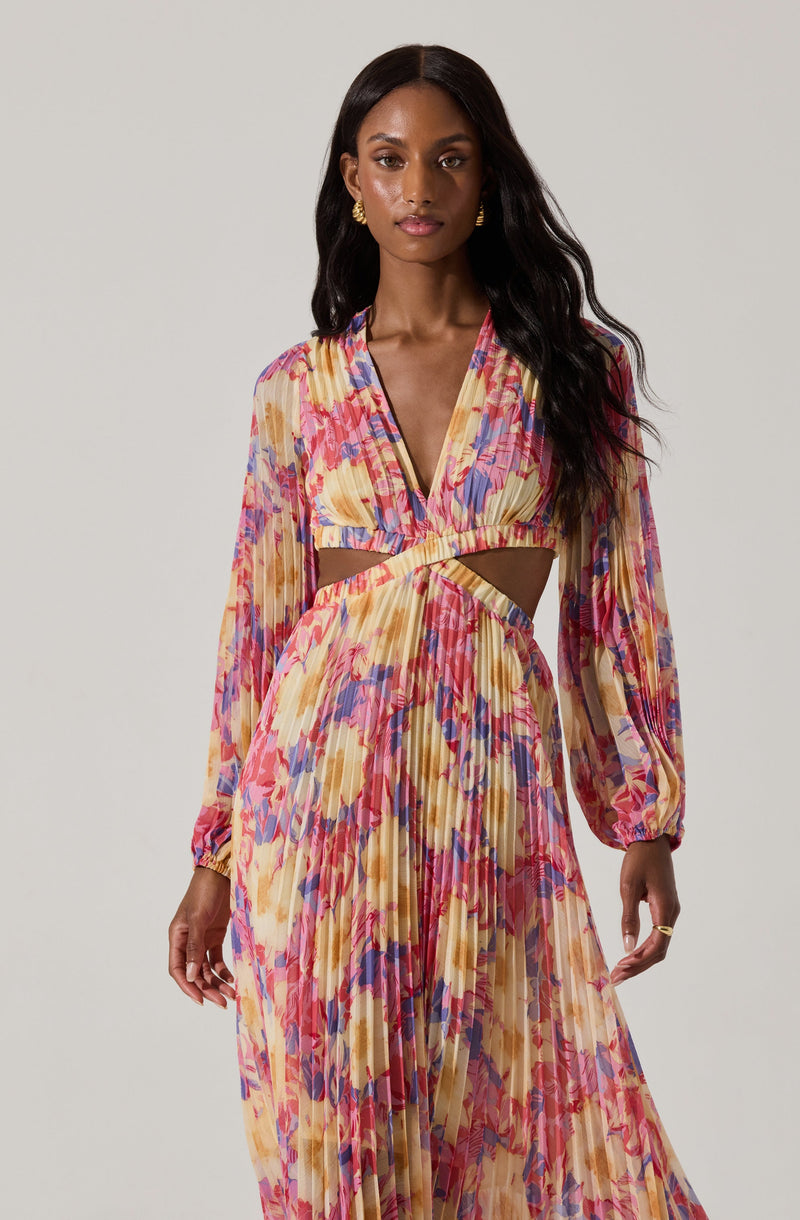 A woman is wearing a colorful, floral-patterned dress with long, puffed sleeves. The dress features a deep V-neckline, a cutout at the waist, and a pleated skirt that extends to the floor. The fabric has a vibrant, abstract floral print in shades of pink, purple, yellow, and beige.