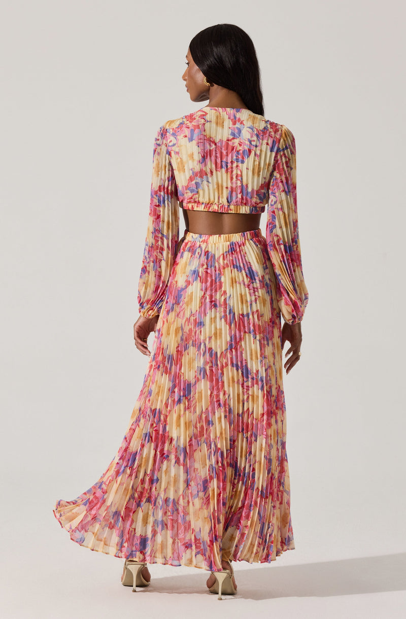 A woman is wearing a two-piece outfit with a colorful, floral, pleated design. The top is a cropped, long-sleeve blouse with puffed sleeves and a ruffled, elasticized neckline. The matching skirt is high-waisted, floor-length, pleated, and features a vibrant pattern of brushstroke-style flowers in shades of pink, purple, yellow, and orange. She is also wearing beige high-heeled shoes.
