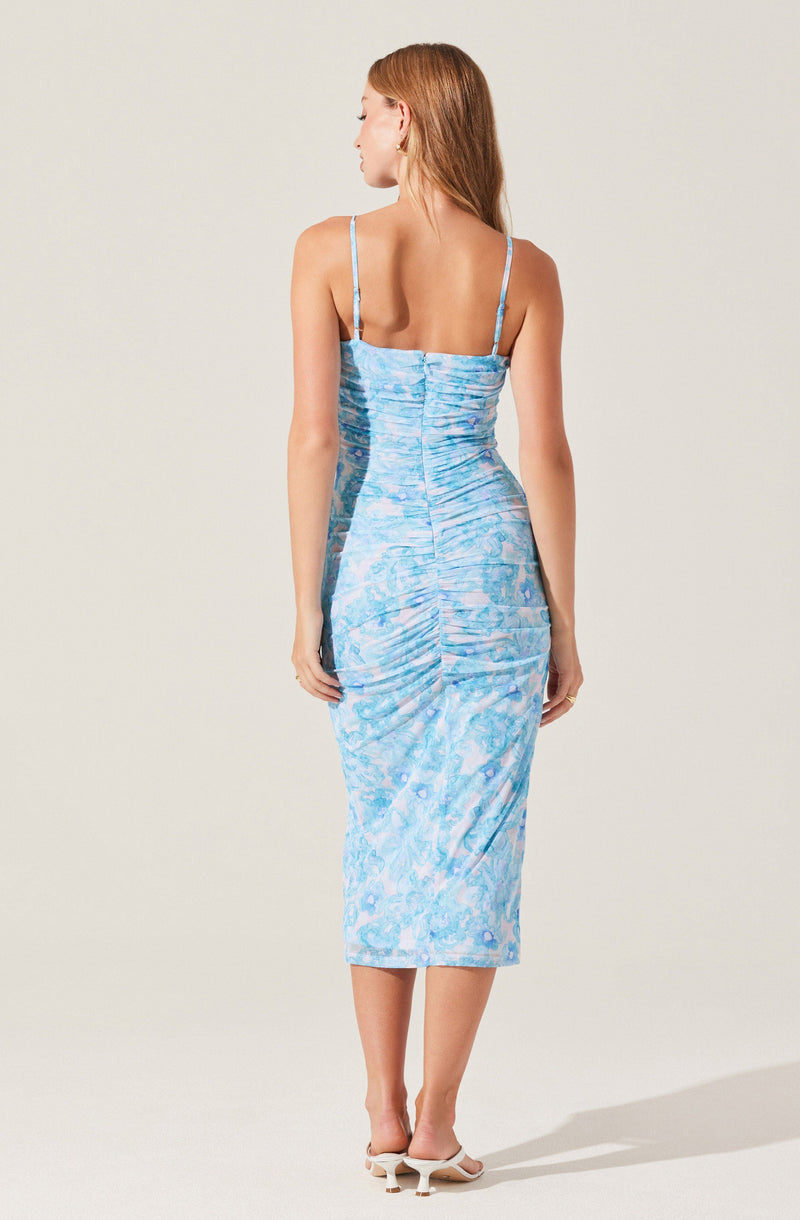 A woman is wearing a fitted, sleeveless, midi-length dress with thin spaghetti straps. The dress has a light blue and darker blue marbled pattern with a ruched detail running vertically down the back. She is also wearing light-colored, open-toed heels.