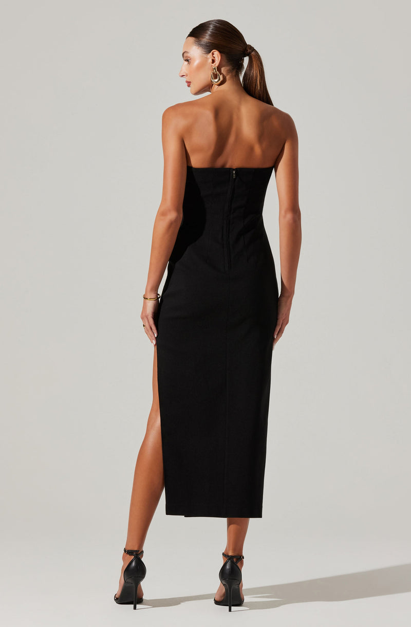 A woman is wearing a strapless, black, full-length dress with a slit on one side. The dress has a fitted silhouette with a back zipper closure. She is also wearing high-heeled sandals and gold jewelry, including hoop earrings, a bracelet, and rings. Her hair is styled in a sleek, low ponytail.