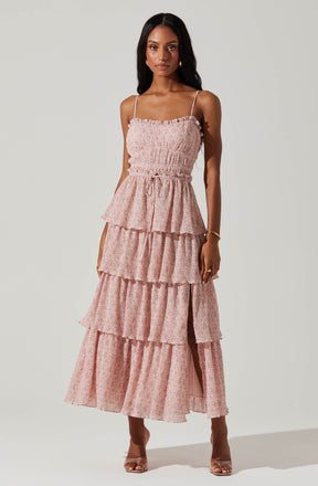 A woman is wearing a light pink, tiered maxi dress with a floral pattern. The dress features thin spaghetti straps, a smocked bodice, and a drawstring at the waist. The skirt has multiple ruffled layers, with a slit showing her leg. She is accessorizing with a gold bracelet, gold earrings, and transparent heeled sandals.