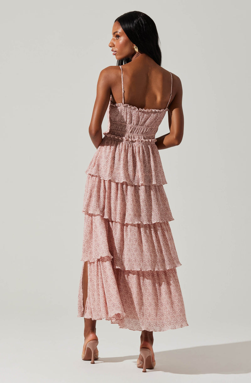A woman is wearing a pink, tiered maxi dress with thin spaghetti straps. The dress has a smocked bodice and ruffled layers that create a flowing silhouette. She has on beige high-heeled shoes and gold earrings.