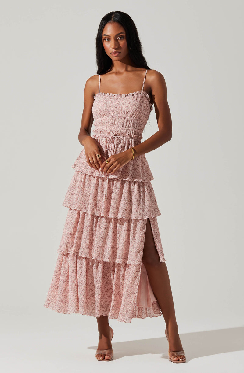 A woman is wearing a pink, tiered, ruffled maxi dress with thin spaghetti straps. The dress features multiple layers with a subtle, small print pattern and a side slit that reveals her leg. She is also wearing clear-strap high-heeled sandals and accessorized with a gold bracelet and rings.