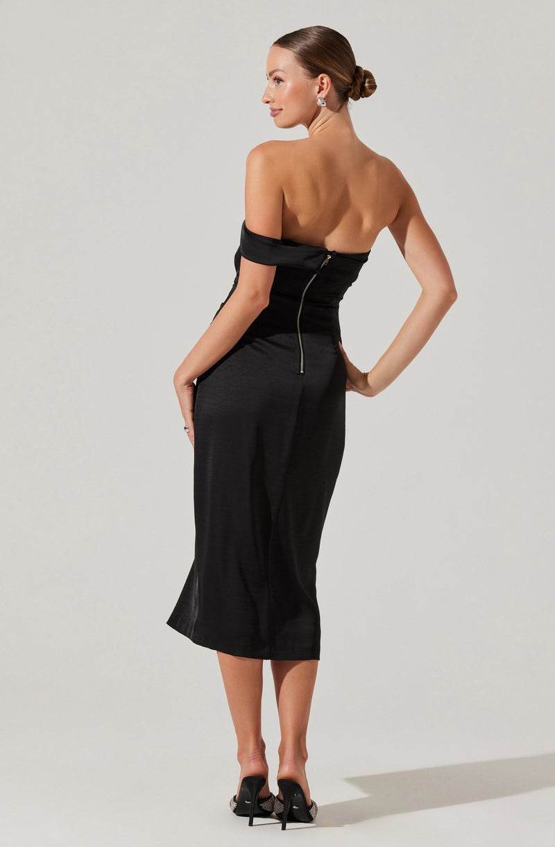 A woman is wearing an off-the-shoulder black dress with a zipper running down the back. The dress is fitted at the waist and hips, and the skirt is midi-length, reaching below the knees. She is also wearing black high-heeled shoes with decorative bows at the back.