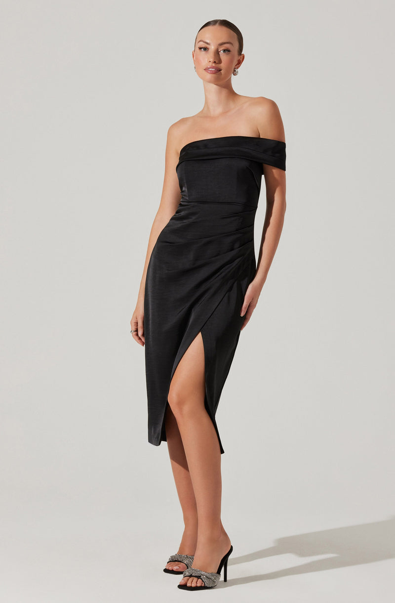 A woman is wearing an elegant off-the-shoulder black satin dress with a fitted bodice. The dress features a draped neckline and a high slit on the front left side, revealing her leg. She pairs the dress with open-toe high heels that have embellished straps.