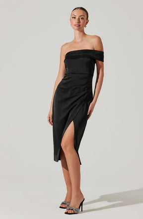 A woman is wearing an elegant off-the-shoulder black satin dress with a fitted bodice. The dress features a draped neckline and a high slit on the front left side, revealing her leg. She pairs the dress with open-toe high heels that have embellished straps.