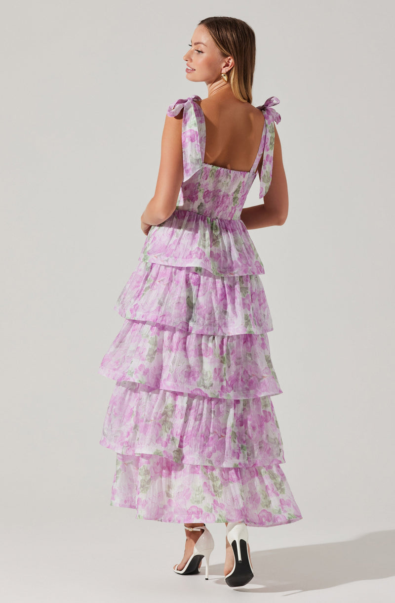 A woman is wearing a sleeveless, tiered maxi dress with shoulder ties. The dress features a purple, pink, and green floral pattern with ruffled layers. She is paired with white high-heeled shoes that have black accents.