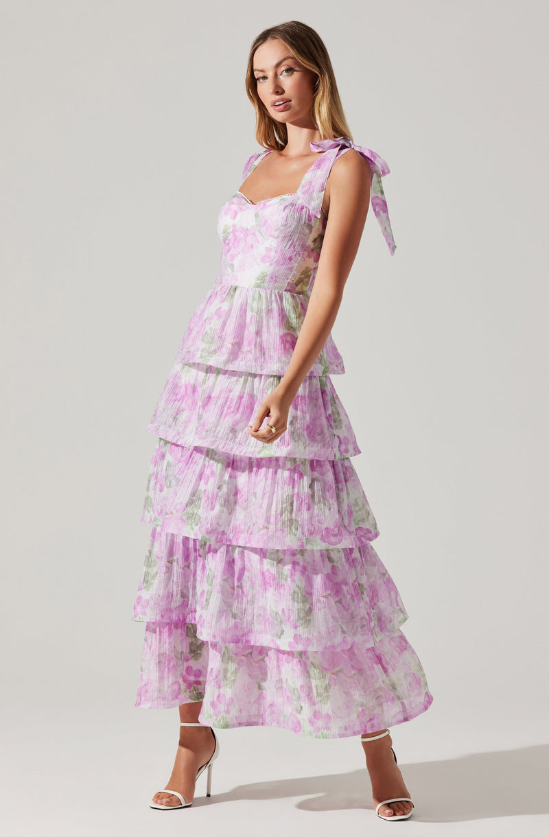 A woman is wearing a sleeveless, floor-length dress with tiered layers. The dress features a light purple and pink watercolor floral pattern and has shoulder straps tied into bows. She is also wearing high-heeled sandals.