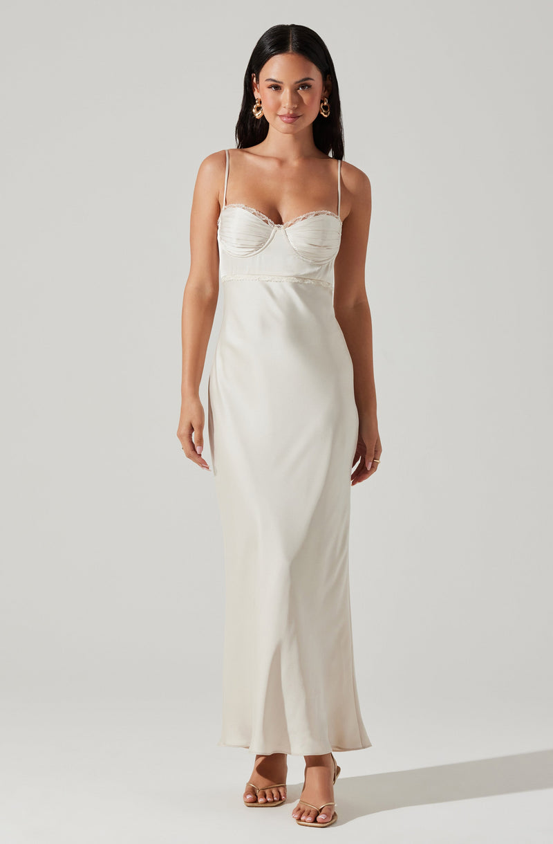 A woman is wearing a sleeveless, off-white satin dress with thin spaghetti straps. The bodice has a gathered design with subtle lace trim along the top edge, and the dress flows down to the floor with a slightly flared hem. She accessorizes with large gold hoop earrings and open-toed high heel sandals.