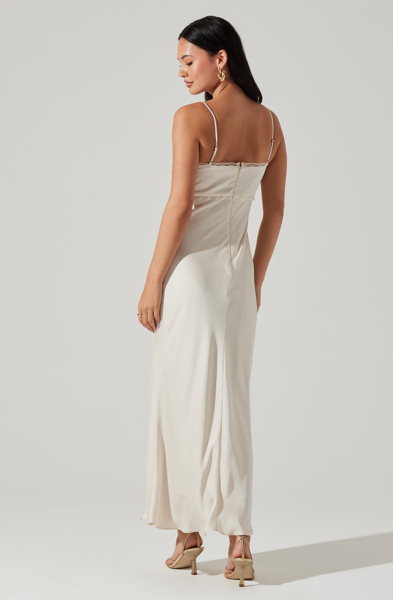 A woman is wearing a long, satin, cream-colored dress with thin spaghetti straps. The dress has a fitted bodice and a flowing, loose-fitting skirt that reaches the floor. She is also wearing nude, high-heeled sandals and large gold hoop earrings.