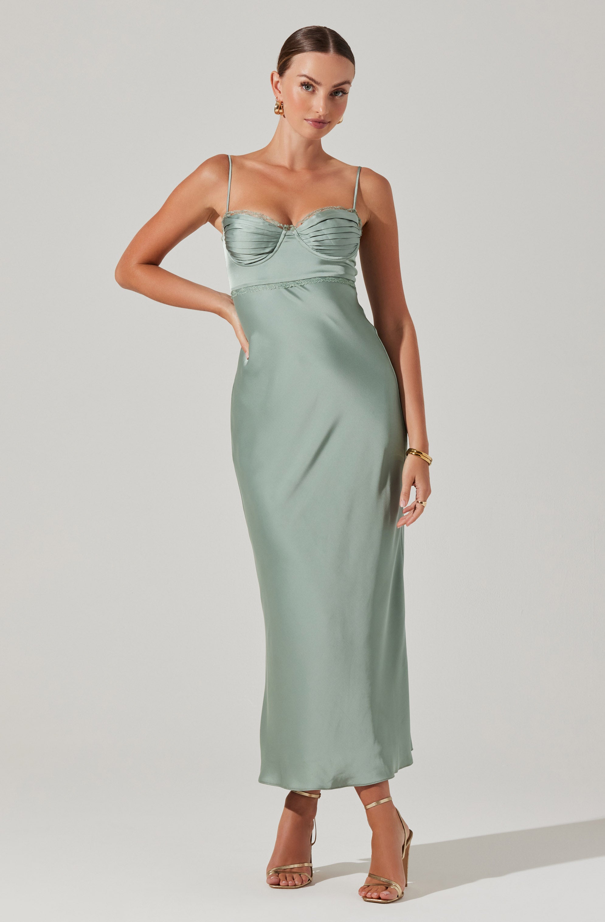 A woman is wearing a long, light green satin dress with thin spaghetti straps. The dress features a ruched, fitted bodice with a sweetheart neckline, and the skirt flows straight down to the floor. She accessorizes with gold jewelry, including earrings and a bracelet, and wears beige high-heeled sandals.