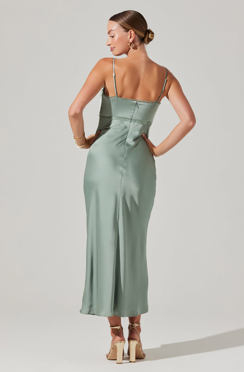 A woman is wearing a light green, satin floor-length dress with thin spaghetti straps. The dress has a fitted bodice with a seam around the waist and a long, flowing skirt with a center slit. She is also wearing beige high-heeled sandals with ankle straps.
