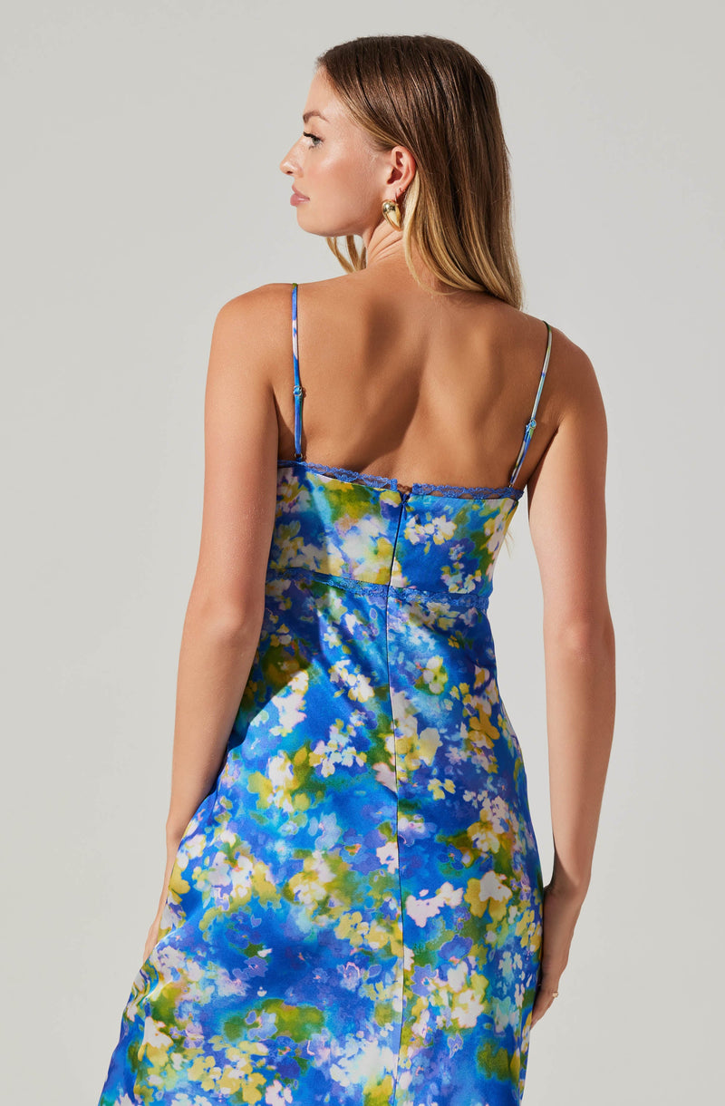 A woman is wearing a sleeveless, silky dress with thin spaghetti straps. The dress features a vibrant, abstract floral pattern in shades of blue, green, yellow, and white. The back of the dress has a visible zipper and a delicate blue lace trim along the top edge. She has long, light brown hair and is accessorized with large gold hoop earrings.