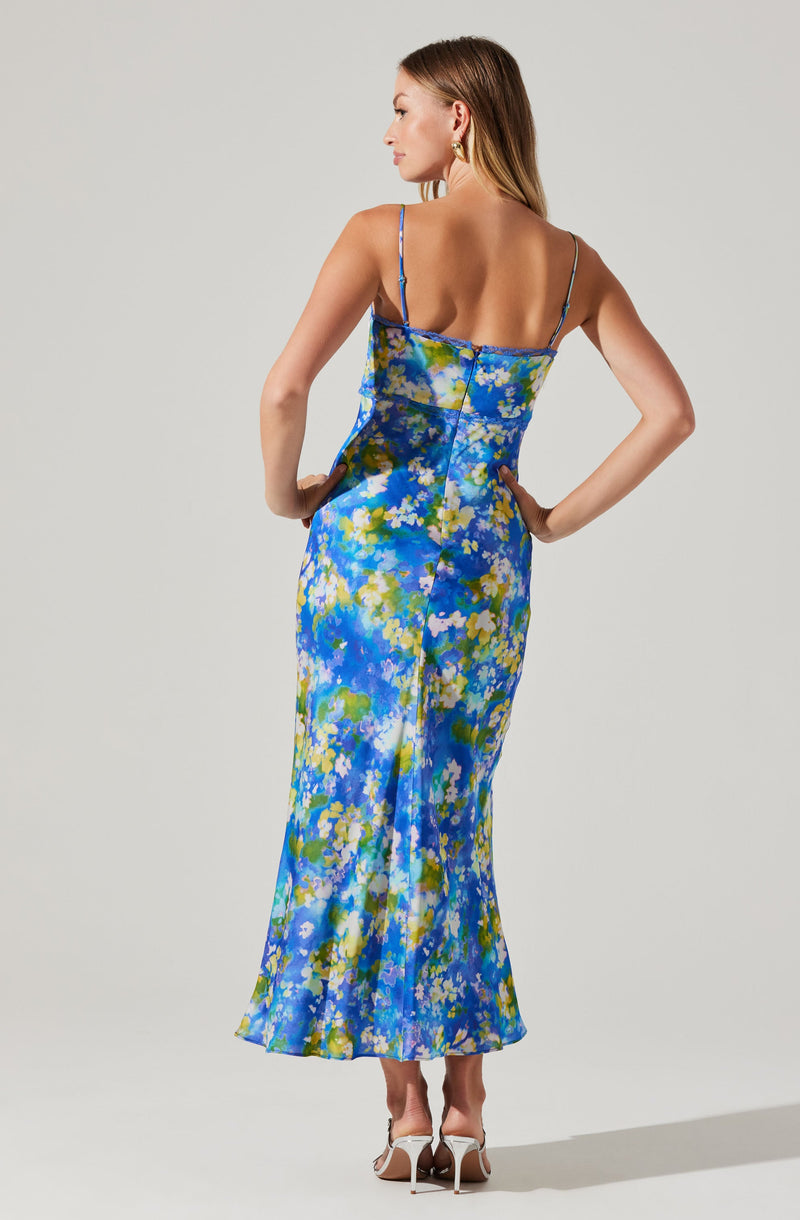 A woman is wearing a sleeveless, ankle-length dress with thin spaghetti straps. The dress features a vibrant, abstract floral pattern in shades of blue, green, yellow, and white.