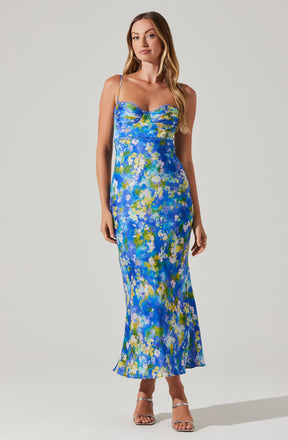A woman is wearing a floor-length, sleeveless dress with thin spaghetti straps. The dress features a colorful, abstract floral pattern primarily in shades of blue, green, yellow, and white. The fabric appears lightweight and flowy. She is also wearing silver, open-toe high-heeled sandals.