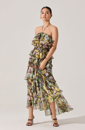 A woman is wearing a strapless, flowy maxi dress with a floral pattern in shades of purple, green, yellow, and brown. The dress features layered ruffles on the bodice and cascading down the length of the skirt. She is also wearing high-heeled sandals with thin ankle straps.