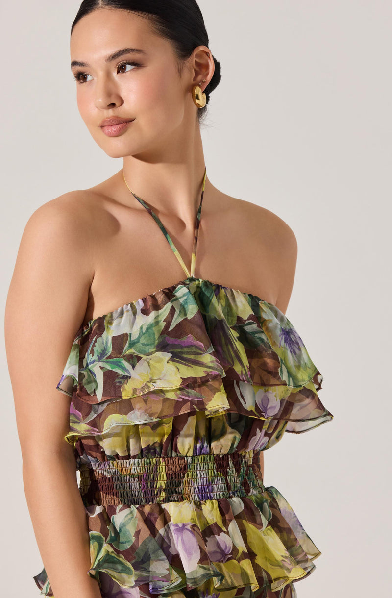 A woman is wearing a sleeveless, strapless dress with a floral pattern featuring shades of green, yellow, purple, and brown. The dress has layered ruffles and a smocked waistband. She accessorizes with large gold hoop earrings and a delicate necklace with a thin chain. Her hair is pulled back into a sleek, low bun.