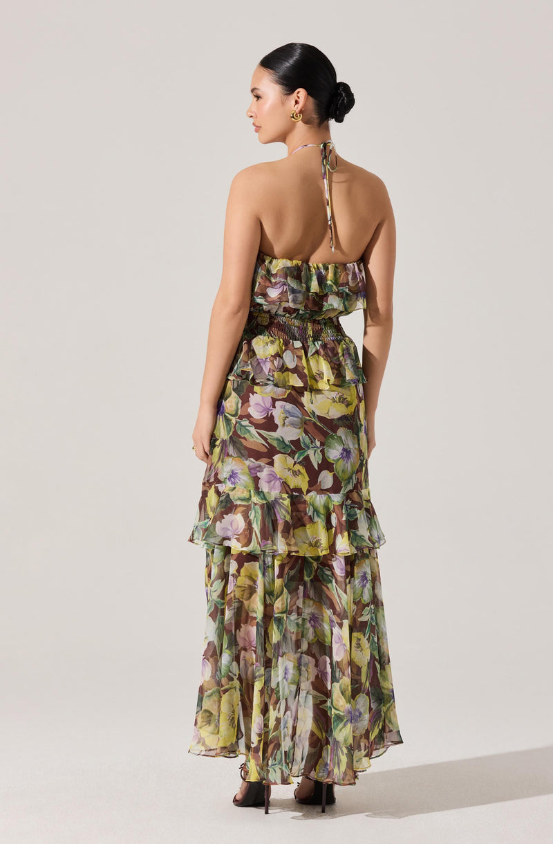 A woman is wearing a long, sleeveless dress with a floral pattern. The dress features a halter neck with a tie at the back, a ruffled top, and multiple tiers of ruffles along the skirt. The fabric is sheer and lightweight with colors including shades of purple, green, yellow, and brown.