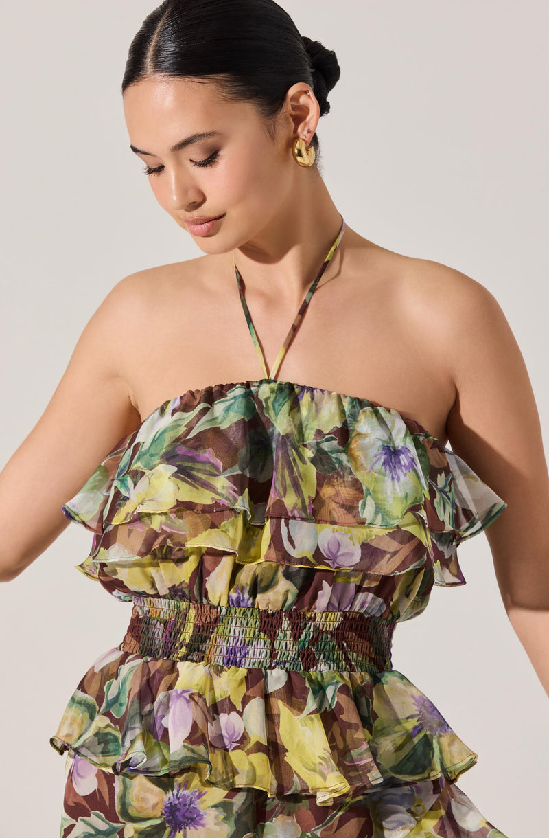 A woman is wearing a strapless, ruffled top with a floral pattern in shades of green, yellow, purple, and brown. The top has layers of ruffles that create a voluminous look. She also has a matching floral skirt with similar ruffles and an elastic waistband.