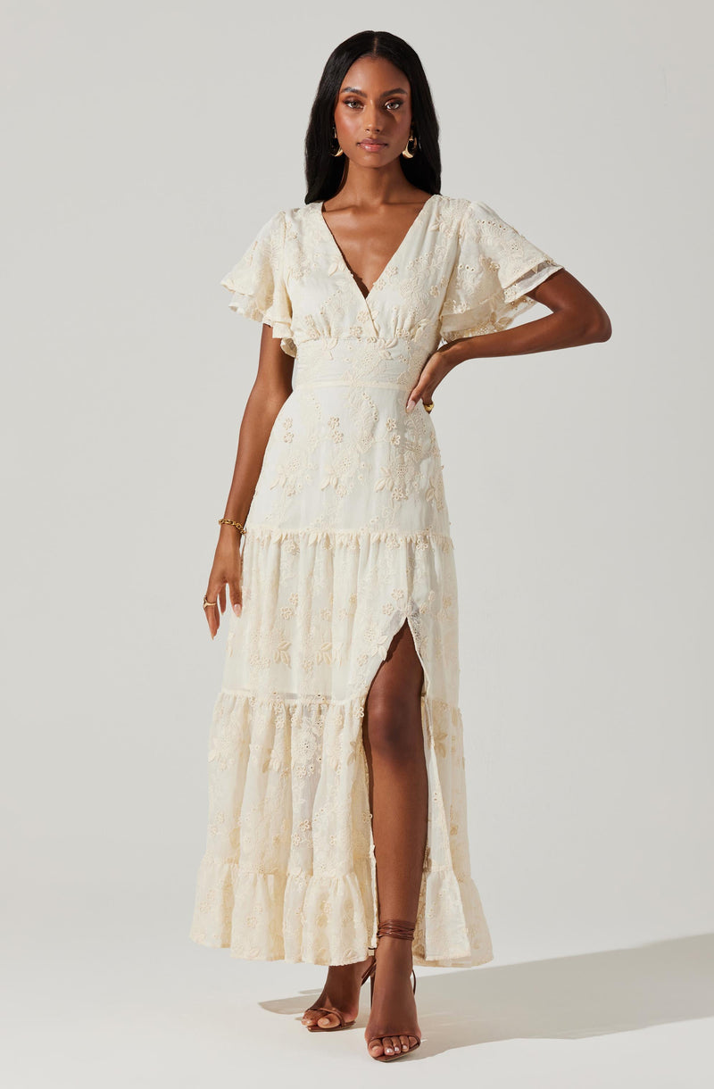 A woman is wearing an off-white, floor-length dress with a deep V-neckline and short, ruffled sleeves. The dress features a high waist, tiered skirt with a slit up the front, and subtle embroidered floral details throughout. She has accessorized with gold earrings, a bracelet, and strappy high heels.