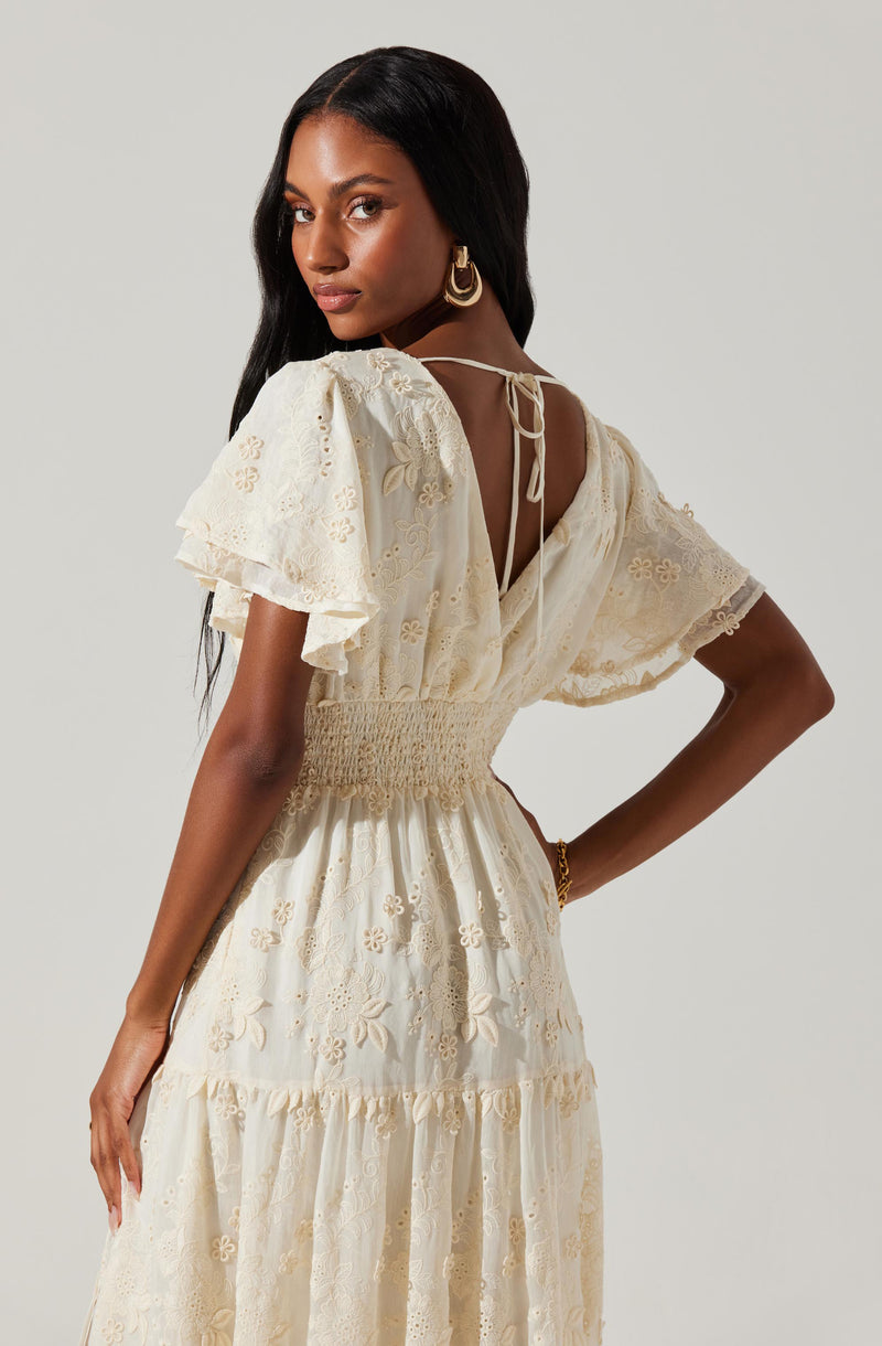A woman is wearing a cream-colored, embroidered dress with short, puffed sleeves and a waistline gathered with smocking. The back of the dress features a tie closure at the neckline. She has long, dark hair and is accessorized with large, statement earrings and a bracelet.