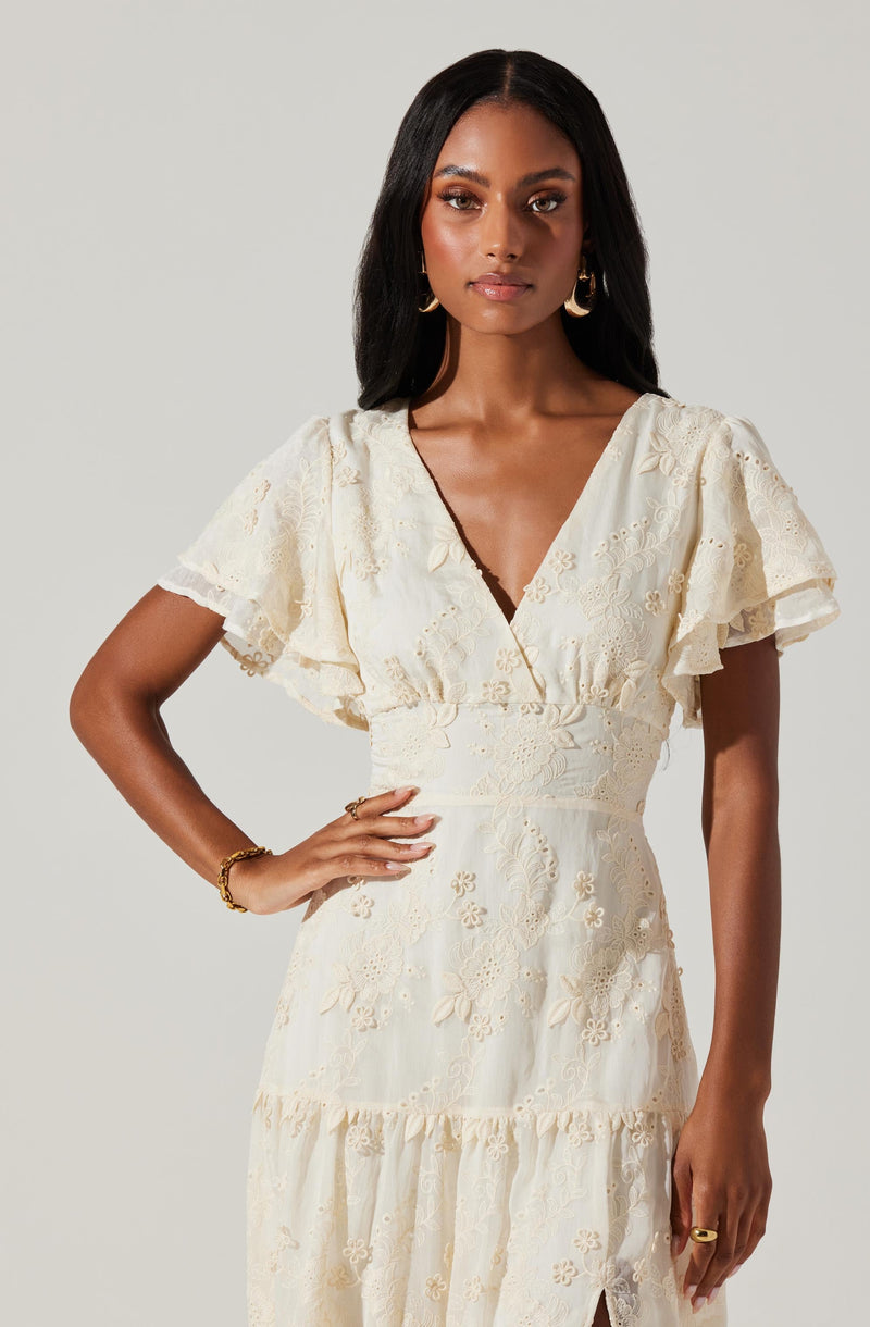A woman is wearing a cream-colored dress with intricate floral embroidery. The dress features a deep V neckline, puffed short sleeves with ruffled edges, a fitted waist, and a tiered skirt with a small slit.