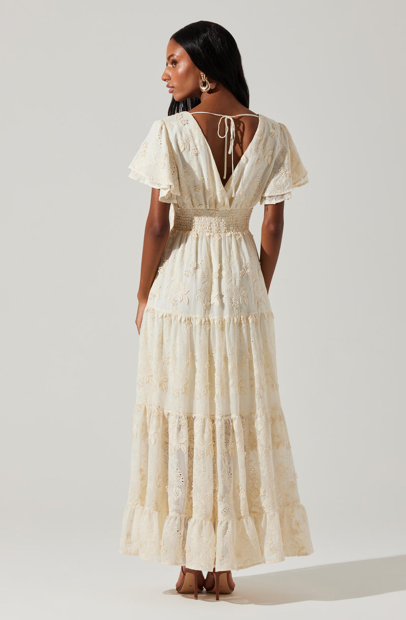 A woman is wearing a long, cream-colored dress with eyelet embroidery. The dress features short, flutter sleeves and a deep V back with a tie closure at the neckline. The waistline is fitted with a smocked detail, and the skirt has a tiered design with multiple gathered sections, creating a flowing and layered appearance. She is also wearing large gold hoop earrings and beige high-heeled shoes.