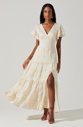 A woman is wearing a long, cream-colored lace dress with a V-neckline and short, ruffled sleeves. The dress has a fitted waist and a tiered skirt with a high slit on the left side, revealing part of her leg. She is accessorized with bracelets, rings, and dangling earrings, and is wearing strappy high-heeled sandals.