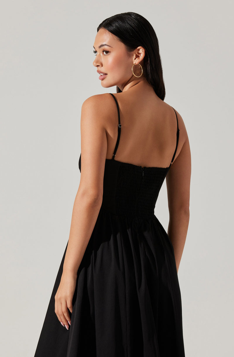 A woman is wearing a black dress with thin spaghetti straps. The dress has a fitted bodice with a smocked or ruched texture and a full, flowing skirt made of shiny, satin-like fabric.